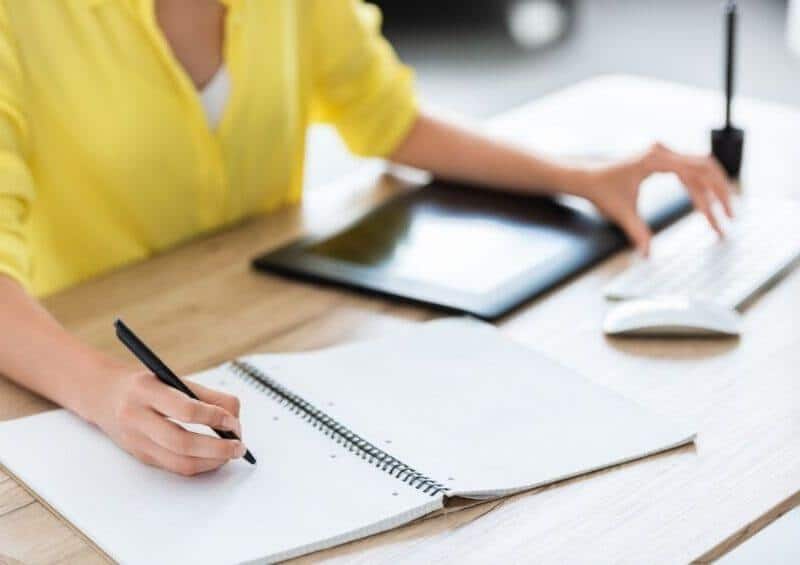 woman wearing a yellow shirt writing on a notepad and typing on keyboard