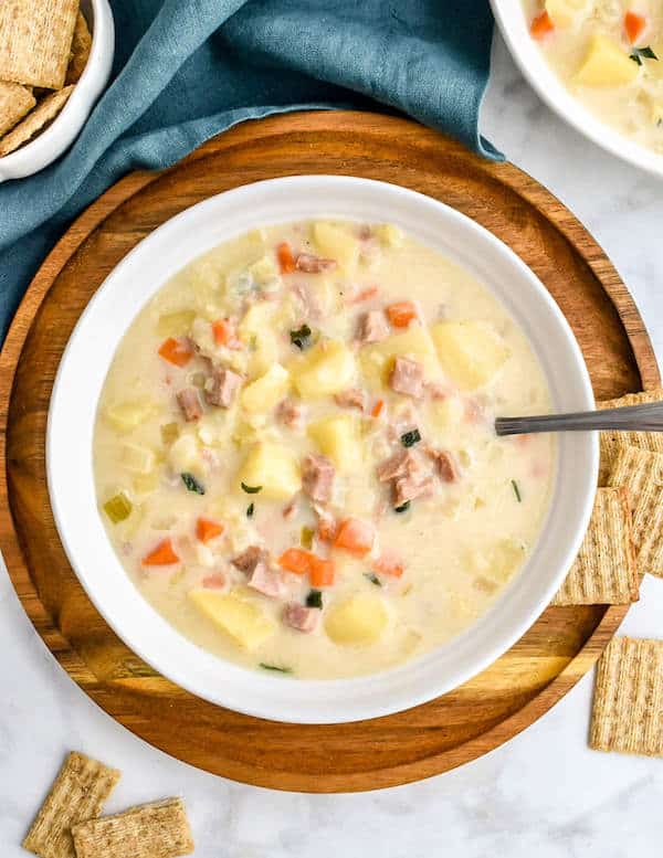 A bowl of Slow Cooker Ham and Potato Soup on a wooden plate next to crackers in a bowl, blue napkin, and on the grey marble countertop.