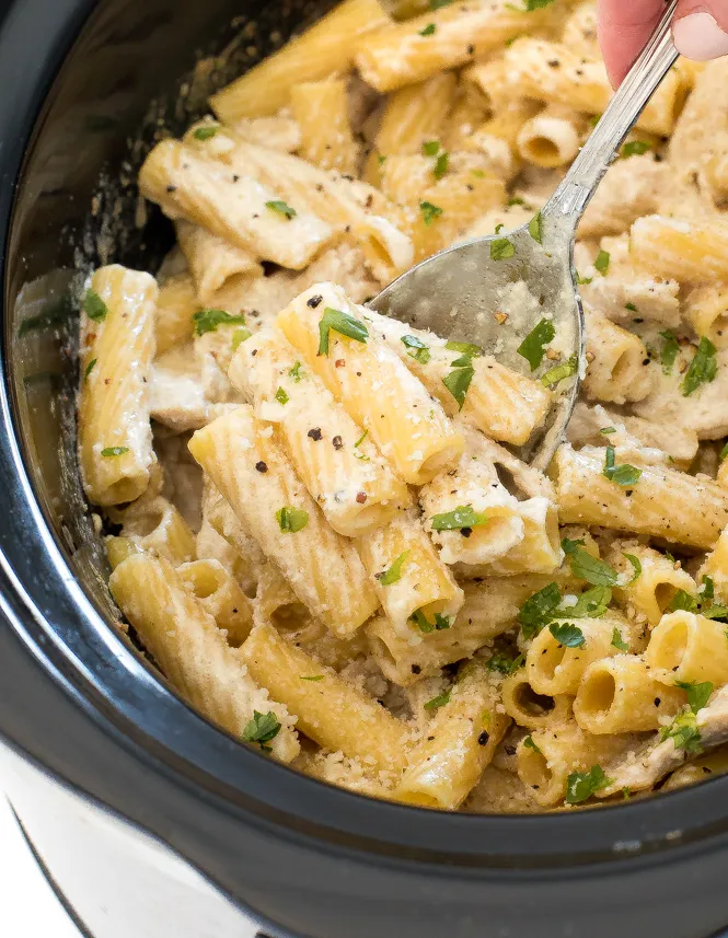 A hand with a metal spoon stirring Chicken Alfredo in a slow cooker