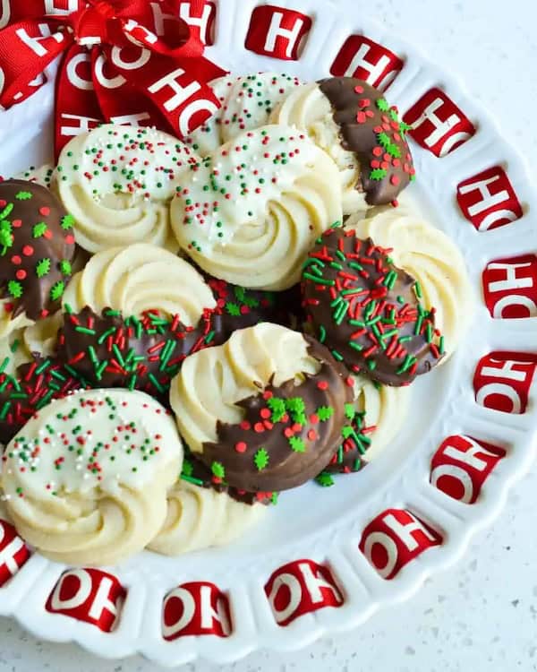 Golden butter cookies piped into swirls, some dipped in white or dark chocolate and topped with red-and-green sprinkles, stacked on a holiday platter.