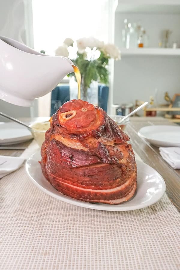 Golden roasted ham on a platter with pouring ham gravy beside it for a festive dinner.