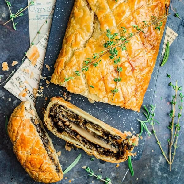 Slice of vegan mushroom wellington with crispy pastry crust and hearty mushroom filling, served on a dark festive board.
