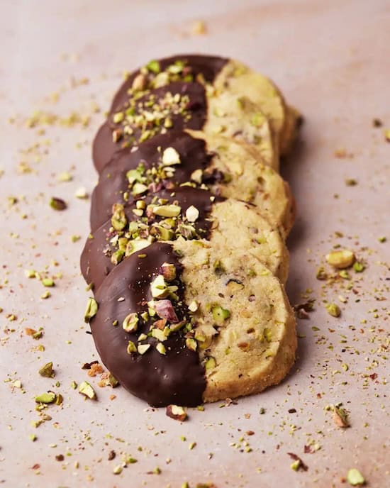 Pistachio shortbread cookies topped with chopped green pistachios and partially dipped in dark chocolate, arranged for a festive holiday Christmas cookie platter.