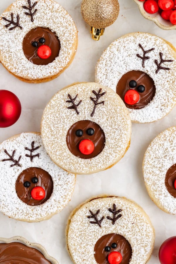 Reindeer Linzer cookies with hazelnut bases, Nutella filling visible through cut-out centers, chocolate-piped antlers and red candy noses, dusted with powdered sugar for a Christmas cookie tray.
