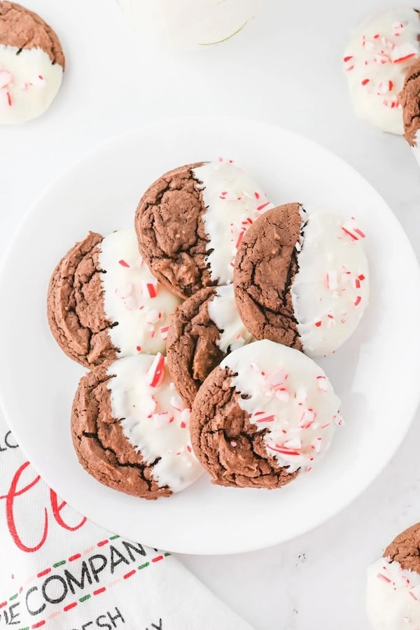 Chocolate cookies half-dipped in white chocolate and sprinkled with red and white crushed candy canes, arranged on a white plate for a Christmas cookie setup.