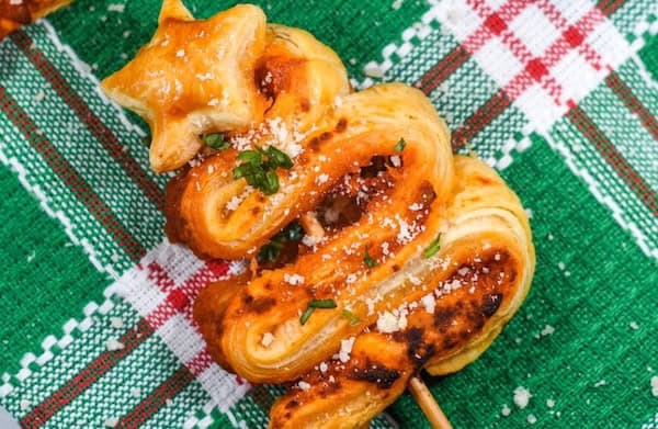Puff pastry Christmas trees sprinkled with herbs on a baking sheet — festive finger food for the holidays.
