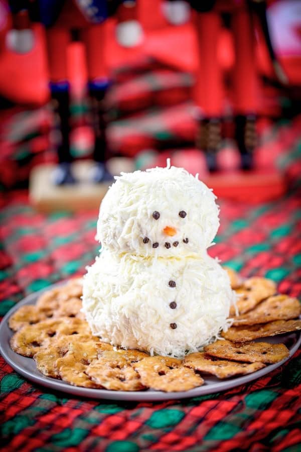 Ranch snowman cheeseball with pretzel arms and carrot nose surrounded by crackers on a festive plate.